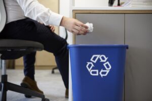 In an office setting, a person skillfully manages waste disposal by tossing a crumpled piece of paper into a blue recycling bin adorned with the white recycling symbol. Seated comfortably, they wear dark pants and brown shoes, demonstrating mindful trash handling practices.
