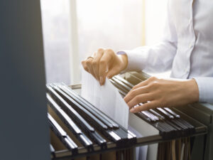 A person in a white shirt is decluttering as they organize documents in a filing cabinet. The open drawer reveals neatly arranged folders. Soft light filters through the window, creating a sense of calm and order in this hazard-free zone.