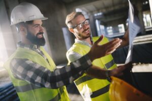 Two construction workers wearing reflective vests and hard hats discuss a blueprint with keen situational awareness at the construction site. One points at the plans, while the other listens attentively. Sunlight filters through the unfinished structure in the background, highlighting their focus on workplace safety.