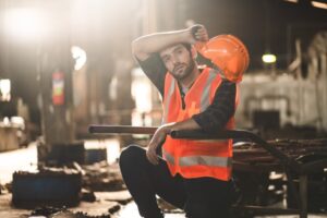 A construction worker in an orange safety vest and helmet looks tired, wiping sweat from his brow. Kneeling in an industrial setting, blurred machinery surrounds him as the warm, atmospheric lighting highlights the science behind heat stress and its effects on the body.