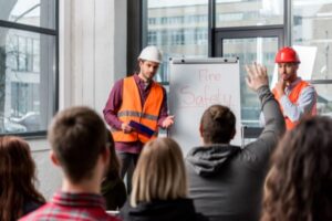 Two men in safety vests and helmets lead a fire safety presentation. One writes on a flip chart about reducing risks, while the other holds a notepad. Several attendees face them, one raising a hand. Large windows in the background let in natural light.