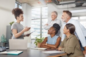 A woman stands at a table holding a notepad, fostering positive communication with four colleagues who are seated and listening intently. The group is in a modern office with large windows in the background, creating an atmosphere that encourages workplace safety and collaboration.