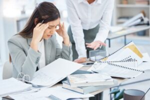 A woman in a business suit sits at a cluttered desk, clearly experiencing workplace anxiety. Holding her head in frustration, she listens as another person standing nearby gestures towards the documents. The scene vividly captures the tension often found in an office setting.