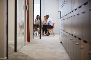 A group of three people are seated around a table having a discussion in a modern office hallway. The hallway is lined with lockers on the right and glass partitions on the left. The space is well-lit and has a contemporary design.