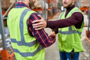 Two men in high-visibility vests are shaking hands in a warehouse, showcasing how to support coworkers. One wears a red plaid shirt, the other a maroon sweater. Shelves stacked with boxes fill the background, setting the scene for collaboration and mutual support.