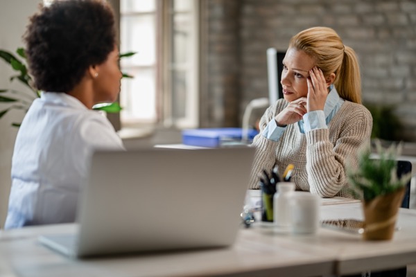 Two people sit at a desk in an office setting. One, wearing a sweater, appears to be expressing frustration or concern. The other person listens attentively. A laptop and potted plant are on the desk. Bright natural light filters through a window.