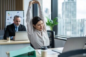 A woman in a light shirt practices workplace safety by stretching with her arms raised at a desk, appearing relaxed. A man works on a laptop in the background. Coffee cups and office supplies are on their desks, with a large window showing city buildings outside.