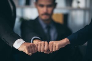 Three people wearing business suits bump fists together in the foreground, symbolizing teamwork and unity, while a man in the background looks on with a serious expression.