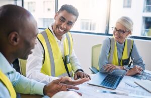 Three people wearing yellow safety vests sit at a table with a laptop and documents, smiling and discussing something in a bright office setting.