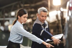 Two people, a young woman in a light blue shirt and an older man with gray hair in a work jacket, stand side by side indoors, pointing and discussing something outside the frame. The background is blurred.