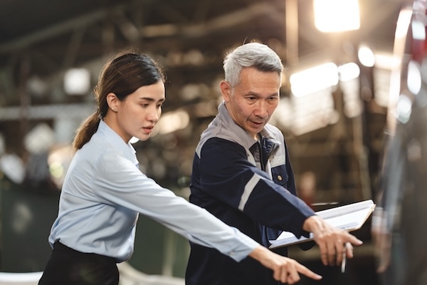 Two people, a young woman in a light blue shirt and an older man with gray hair in a work jacket, stand side by side indoors, pointing and discussing something outside the frame. The background is blurred.