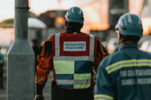 Two people in safety gear stand outdoors. One, wearing a helmet and vest labeled "Coastguard Rescue Officer in Charge," faces away, while the other stands nearby. The background is blurred.
