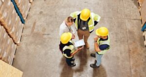 Four people, three wearing yellow hard hats and reflective vests, and one in business attire, stand in a warehouse having a discussion while looking at a tablet. Stacks of boxes are visible around them.
