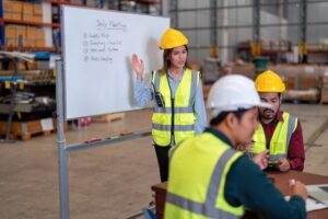 A woman in a safety vest and hard hat leads a daily meeting by a whiteboard with notes, while two men in safety gear listen at a table in a warehouse.