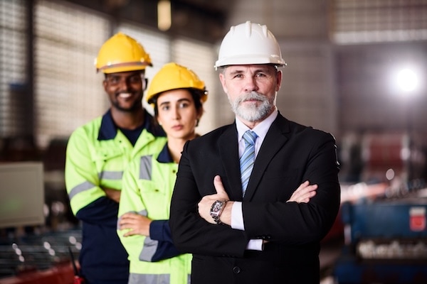 Three people in a factory: a man in a suit and white hard hat stands confidently in front, while two workers in yellow hard hats and safety jackets stand behind him, smiling. Industrial equipment is visible in the background.