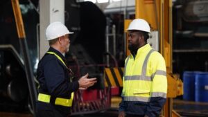 Two workers wearing safety helmets and high-visibility jackets are having a discussion inside an industrial facility, with machinery and equipment visible in the background.