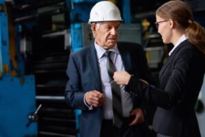 An older man in a suit and white hard hat talks to a woman in a blazer and glasses inside an industrial facility, with large machinery visible in the background.