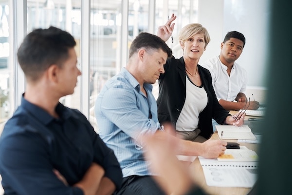 Four people sit at a conference table in a bright office. One woman is raising her hand to speak, while the others listen attentively and take notes. Notebooks and papers are on the table.