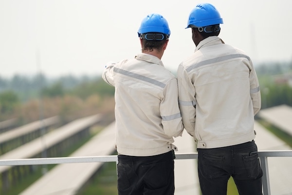 Two workers wearing blue helmets and light-colored jackets stand on a platform, facing away from the camera. One worker is pointing outward, overlooking what appears to be a solar panel field.
