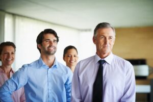 Four professionally dressed people stand indoors, smiling and looking slightly to the right. The group includes both men and women, suggesting a business or teamwork setting in a modern office environment.