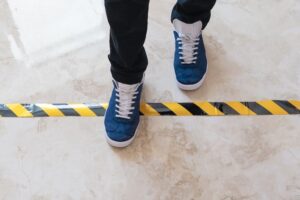 A person wearing blue sneakers stands with one foot over a black and yellow caution tape line on a marble floor.