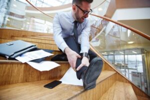 A man in business attire sits on wooden stairs, holding his ankle in pain. Scattered papers, a phone, and a briefcase lie beside him, suggesting he slipped and fell.