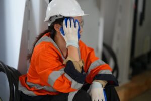 A person in a white hard hat, safety glasses, orange reflective jacket, and gloves sits and rests their head on one gloved hand, appearing tired or thoughtful at a worksite.