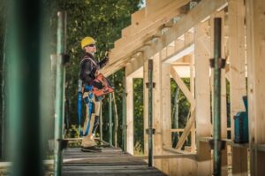 A construction worker wearing a yellow hard hat and safety harness stands on scaffolding while working on the wooden frame of a building under construction, surrounded by trees.