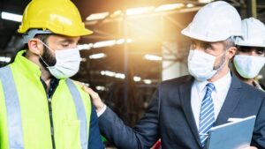 A man in a suit and hard hat talks to a construction worker in a high-visibility vest and helmet. Both are wearing face masks, standing in an industrial setting with another worker visible in the background.
