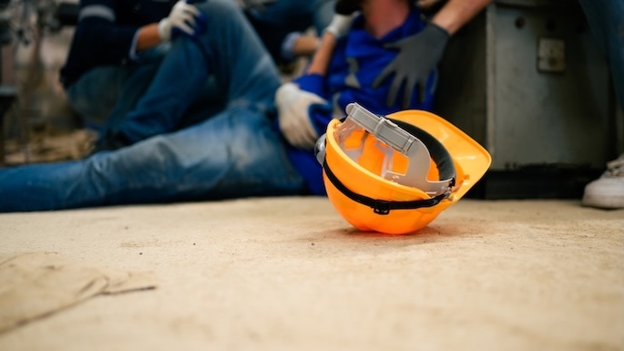 An orange safety helmet lies on the ground in the foreground, while two workers in blue clothing and gloves assist an injured colleague in the blurred background.