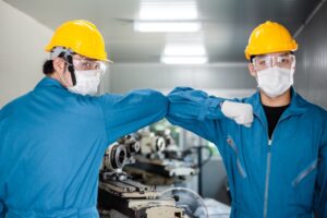 Two factory workers wearing yellow helmets, face masks, safety goggles, and blue coveralls greet each other by touching elbows inside an industrial workshop.