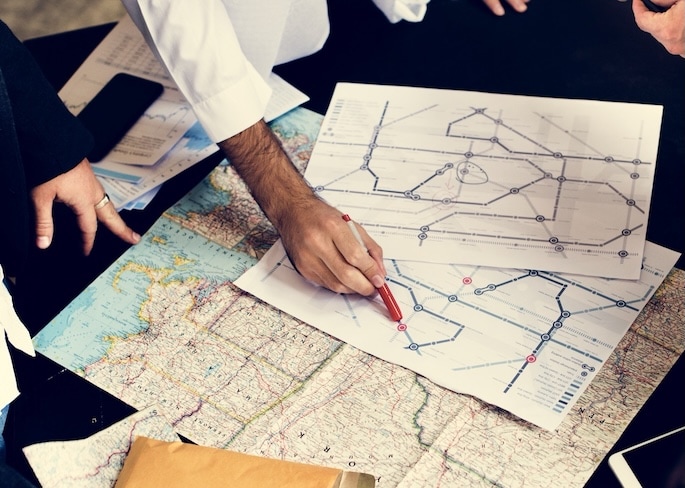 Three people are gathered around a table looking at a map and a transit diagram. One person points at a location on the transit diagram with a red pen, while others observe and discuss.