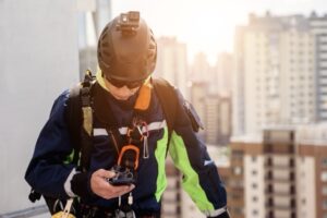 A construction worker wearing safety gear and a helmet with a camera looks at a device in his hand while standing on a rooftop with tall city buildings blurred in the background.