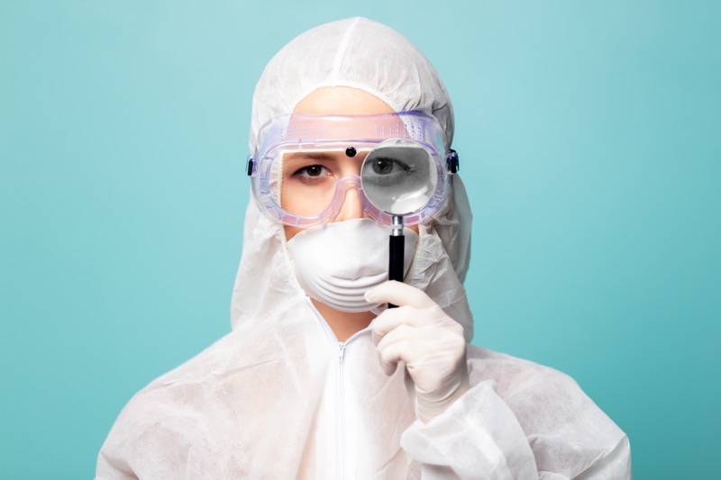Person wearing a protective suit, mask, gloves, and safety goggles holds a magnifying glass up to one eye, enlarging it. The background is a solid light blue.