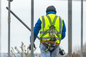 A construction worker in a high-visibility vest and safety harness stands among metal scaffolding, facing away from the camera. The sky is overcast and trees are visible in the background.