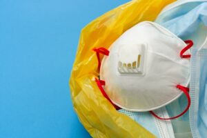 A white protective mask with red straps and a valve is placed on top of blue surgical masks inside a yellow plastic bag, all set against a blue background.