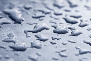 Close-up of water droplets scattered on a smooth, metallic blue surface, with soft lighting creating reflections and a slightly blurred background.