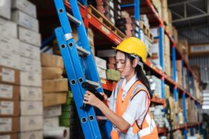 A woman in a hard hat and safety vest uses a barcode scanner while standing on a ladder in a warehouse filled with shelves of boxes.