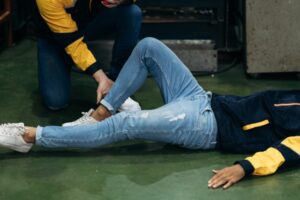A person in jeans and a jacket is lying on the floor while another person kneels beside them, checking or supporting their ankle, possibly providing first aid.