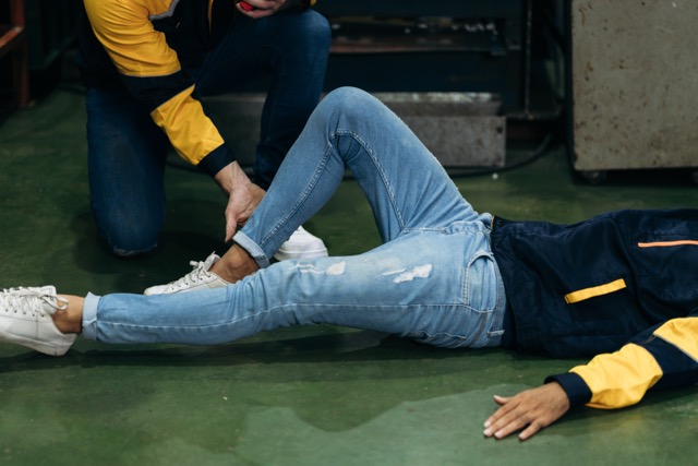 A person in jeans and a jacket is lying on the floor while another person kneels beside them, checking or supporting their ankle, possibly providing first aid.