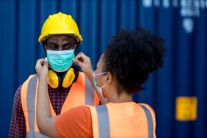 A woman in a safety vest helps a man in a hard hat and safety vest adjust his face mask. The man is also wearing ear protection around his neck, and they are standing in front of a blue background.