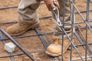 A construction worker wearing tan boots and khaki pants uses pliers to cut a steel rebar on a construction site with sand and steel rods visible on the ground.