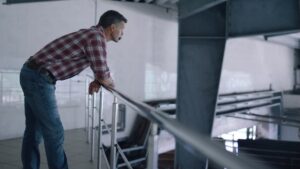 A man in a plaid shirt and jeans leans on a railing, looking thoughtfully into an industrial or warehouse space with metal beams and white walls.