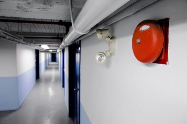 A long, empty hallway with white and blue walls features a prominently mounted red fire alarm for emergency evacuation and a small, attached white light. Doors line the walls, leading to different rooms, and overhead piping is visible on the ceiling.