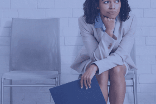 A woman with curly hair, dressed in a light-colored business suit, sits on a chair against a white brick wall. Holding a folder and resting her chin on her hand, she appears deep in thought, likely pondering human resources decisions. An empty chair is next to her.