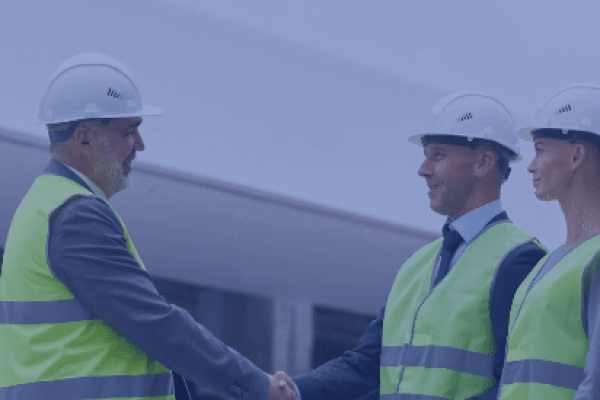 Three people wearing hard hats and neon yellow safety vests are standing indoors. Two of them, a man and a woman holding blueprints, are observing while a third man and the man with blueprints are shaking hands, possibly sealing a deal or agreement overseen by human resources.