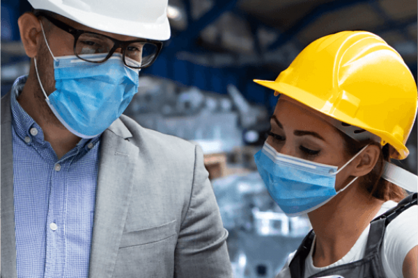 A man in a gray suit and white hard hat, and a woman in a yellow hard hat and safety vest, both wearing blue masks, collaborate about safety in an industrial setting. The man holds a tablet, and the woman holds a clipboard as they discuss work.