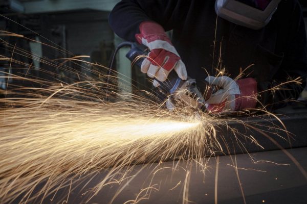 Person wearing protective gloves and face shield uses a handheld angle grinder, producing a burst of bright sparks while cutting through metal in an industrial workspace. The dramatic lighting accentuates the flying sparks, underscoring the importance of grinders safety in such an intense work environment.