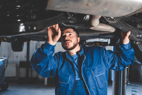 A mechanic in a blue jumpsuit examines the underside of a vehicle raised on an auto lift in a garage. He appears focused as he inspects the components above him. The background shows a workshop environment with various tools and equipment.