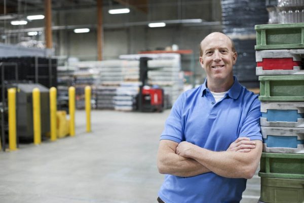 A bald man in a blue polo shirt stands smiling with arms crossed in a large manufacturing plant. He is leaning against a stack of plastic crates. The background shows shelves filled with various materials and equipment.